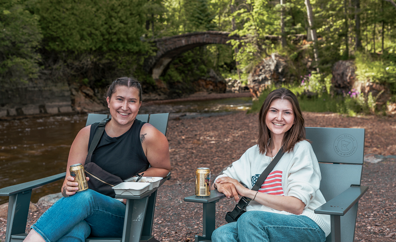 two women seated in front of stone arch bridge with beer; a green foodtruck with container of food; ladies seated on the pier.