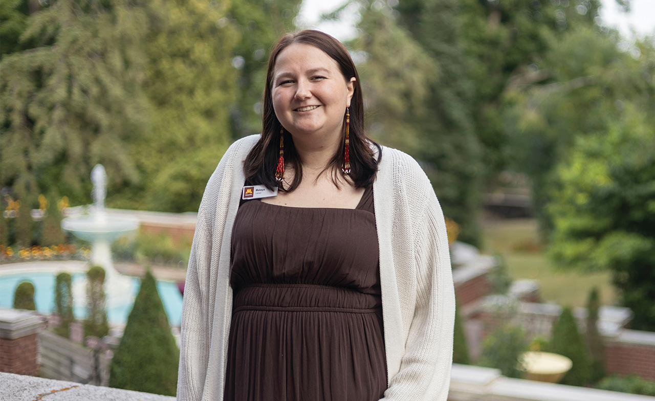 girl in brown dress with fountain in and green trees in the background.
