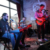 Adam Herman & the Dynamite Club, three men playing in a band in a brick building.