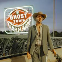 Clayton Ryan, man with hat and suit, lit up sign behind him that reads 'ghost town'