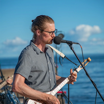 Climes, man playing a string instrument and singing in a mic outside on a lake.