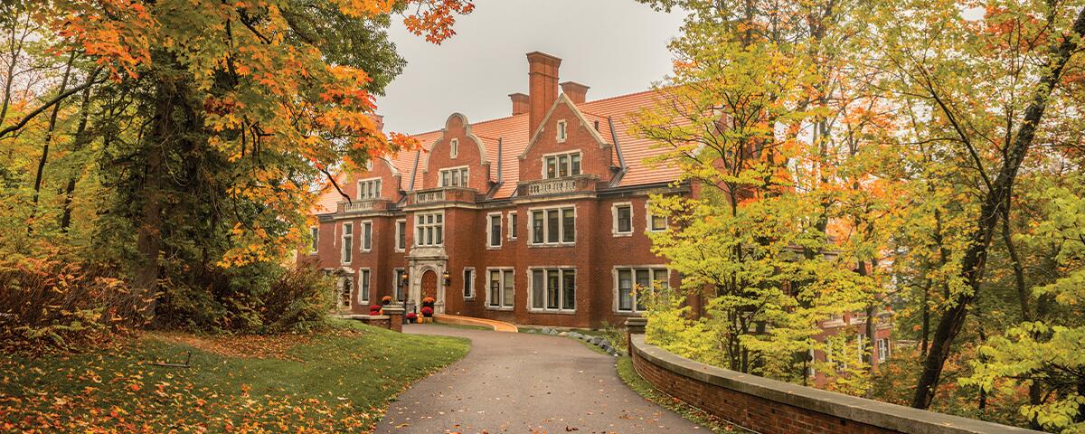 Front of a brick exterior mansion with a curved driveway and colorful fall trees.