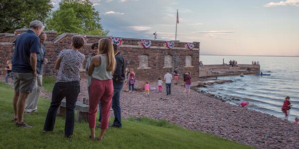 Group of adults stands in a circle chatting on the lawn, boathouse and lakeshore in the background.