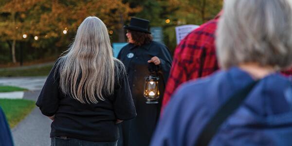 Tour guide in a cloak, holding a lantern and guiding a group of guests down a path.