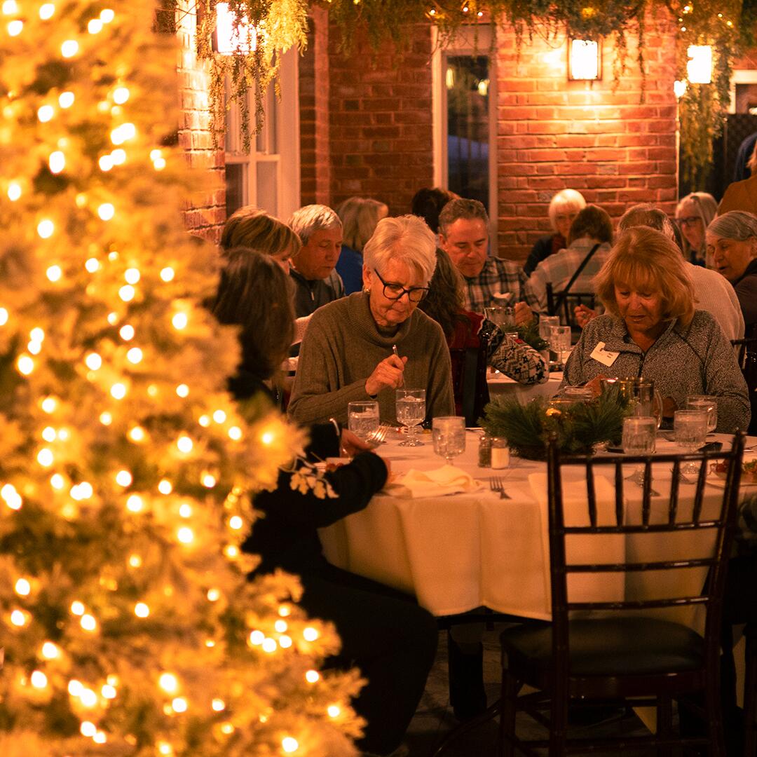 Group of mature women eating at a table, christmas tree lights to the left.