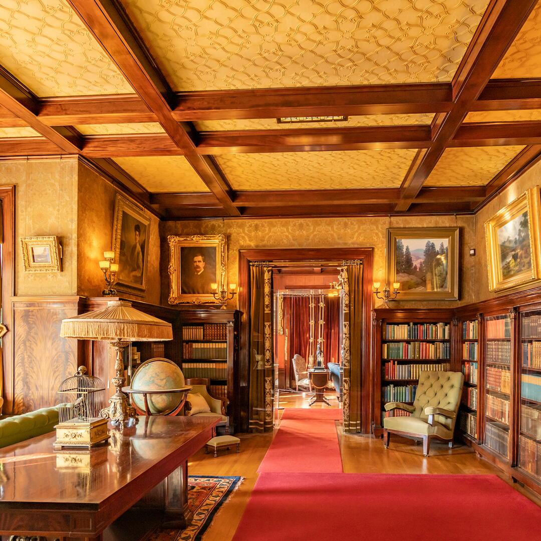 Glensheen library: room with book shelves, long wooden table with a lamp and seating area, and textured wood panel ceiling.