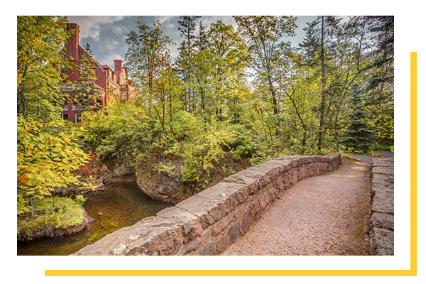 Stone Arch Bridge over Tischer Creek with green trees and the side of Glensheen Mansion in the background.