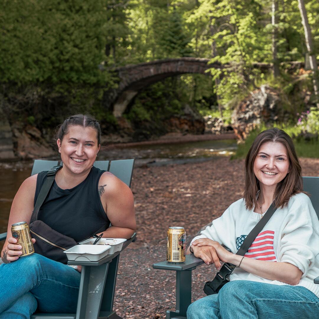 two women seated in front of stone arch bridge with beer; a green foodtruck with container of food; ladies seated on the pier.