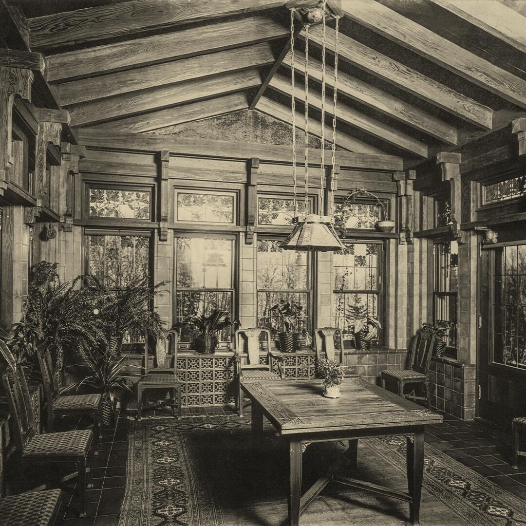 wood table in center of stained glass and wood paneled room, chairs on outside along with plants, light hanging from ceiling.