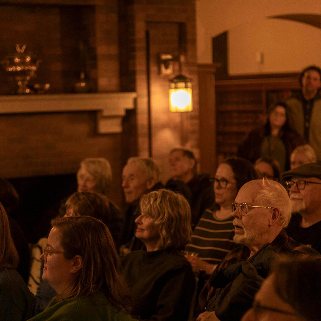 crowd of adults seated and watching a speaker present, brick fireplace in the background.