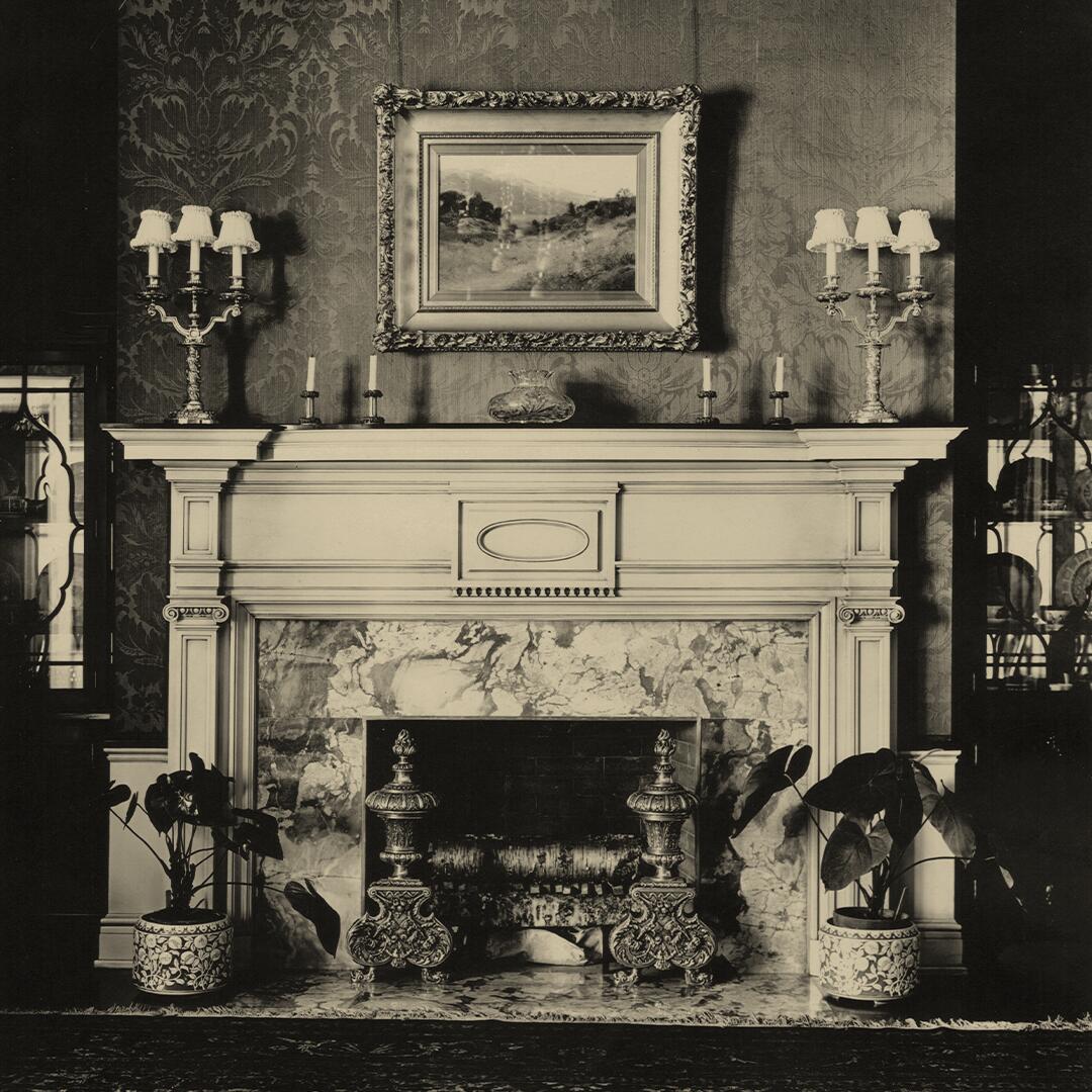 marble and wood fireplace with candelabras, plants below, and oil painting above.