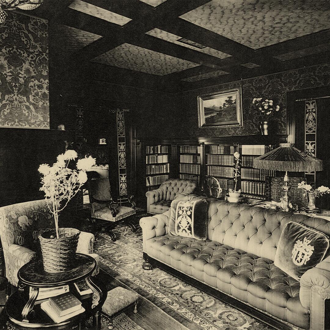 chair and sofa with shelves of books, wood paneled walls and ceiling, and a table with a large lamp.