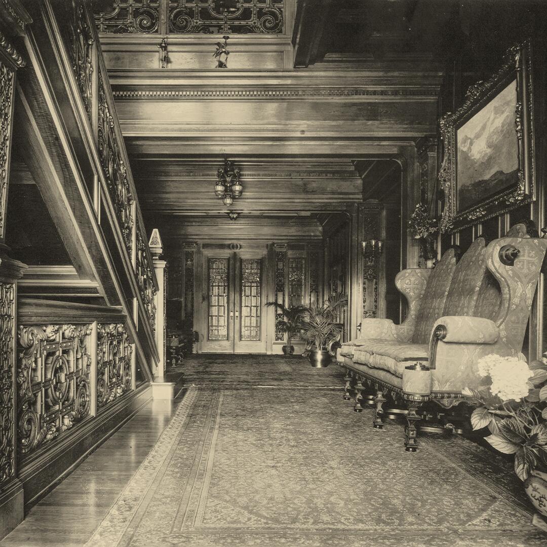 wood paneled hall with wood floors and intricate rug, wood carved stair railing to left and furnishing to the right.