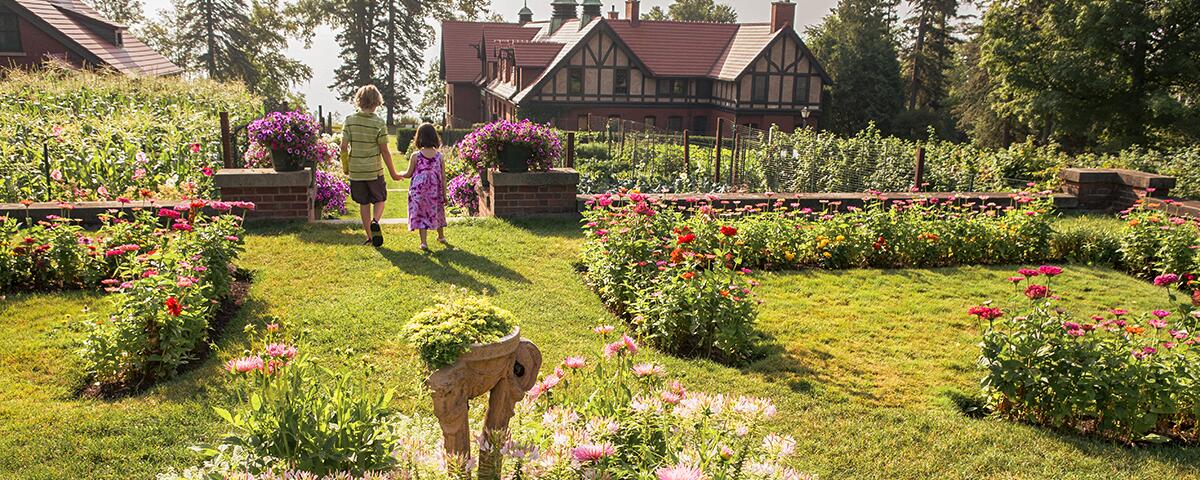 two kids holding hands and walking through grassy garden filled with flowers, bushes, orange carriage house in the background.