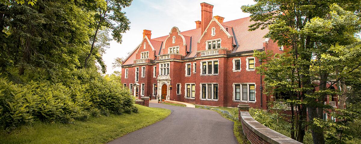 Front of a brick exterior mansion with a curved driveway and green trees.