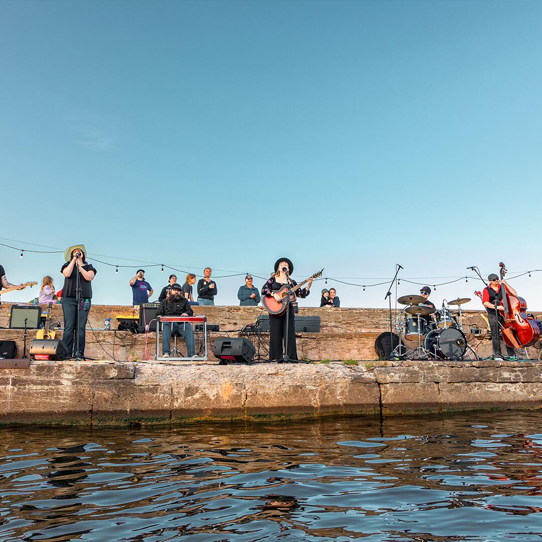 band of singers, a drummer, bassist, and pianist performing on the pier on the lake.