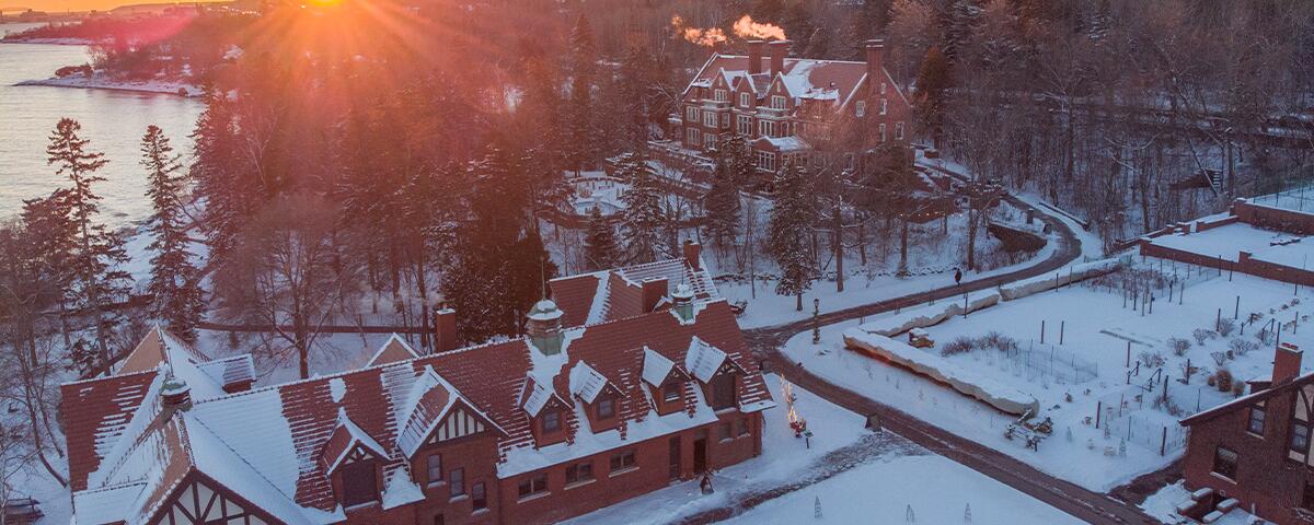 large orange brick carriage house building with the mansion in the back, sun peeking through, snow on the ground.