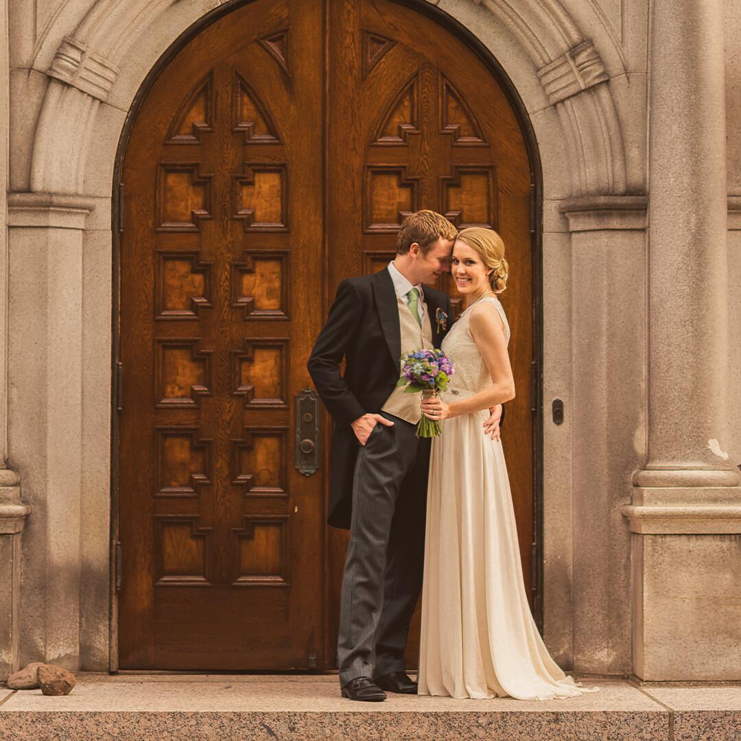 groom and bride posing in front of the tall rounded wooden front doors of the mansion.