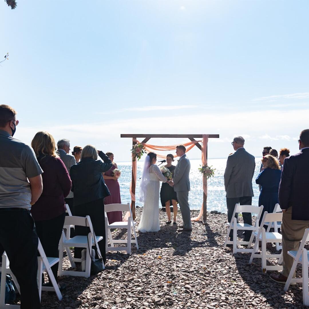 Guests standing and watching a bride and groom join hands at the altar on the stone shores of the lake.
