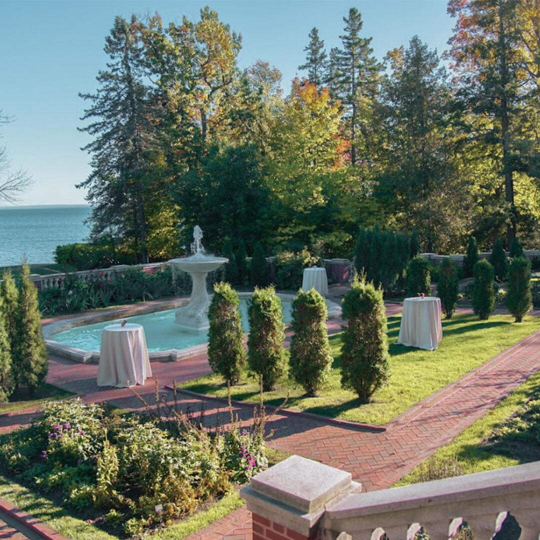 Outdoor formal garden with pruned bushes, grassy areas, and a large fountain, cocktail tables around.