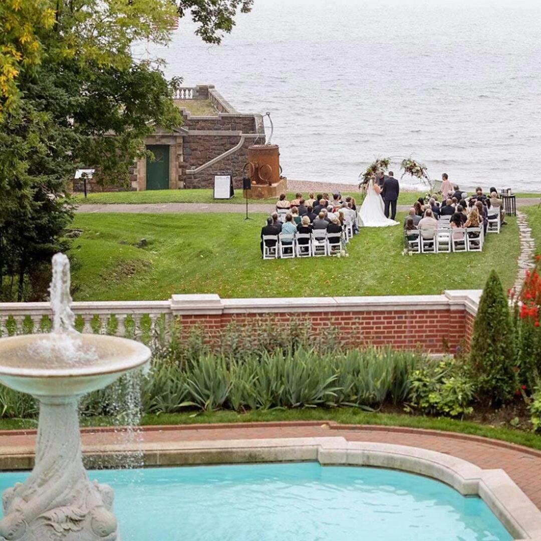 Large fountain in the foreground, overlooking a wedding ceremony on the grassy lawn by the lake shore.