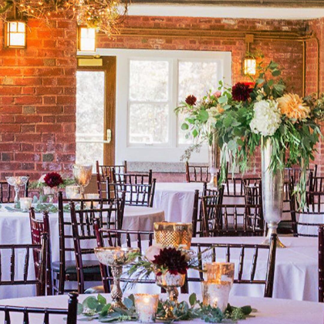 Long brick room filled with white table clothed round tables and chairs, flowers as centerpieces.