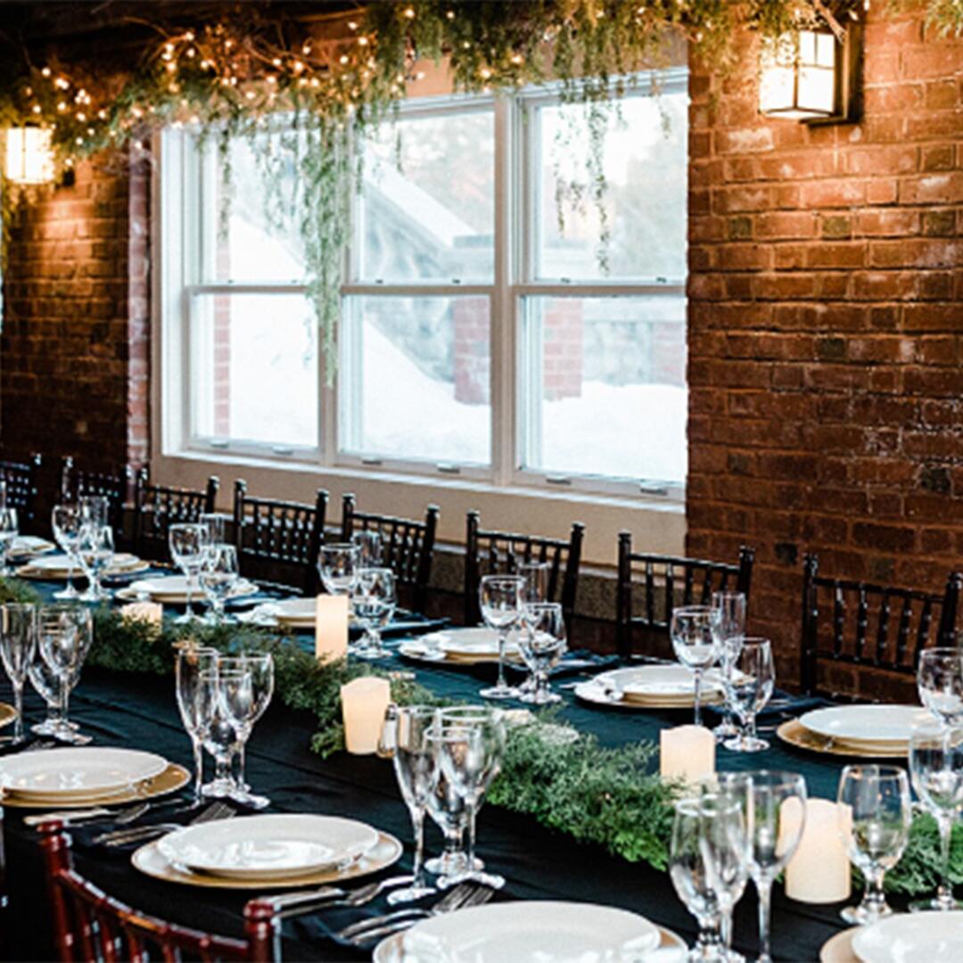 long black table clothed table with plates and glasses set for a dinner in a brick exterior room.