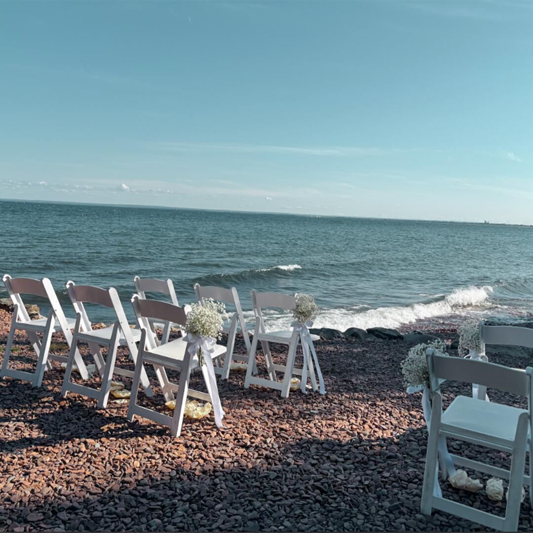 rows of white folding chairs set up on pebble shore of Lake Superior for a wedding ceremony.