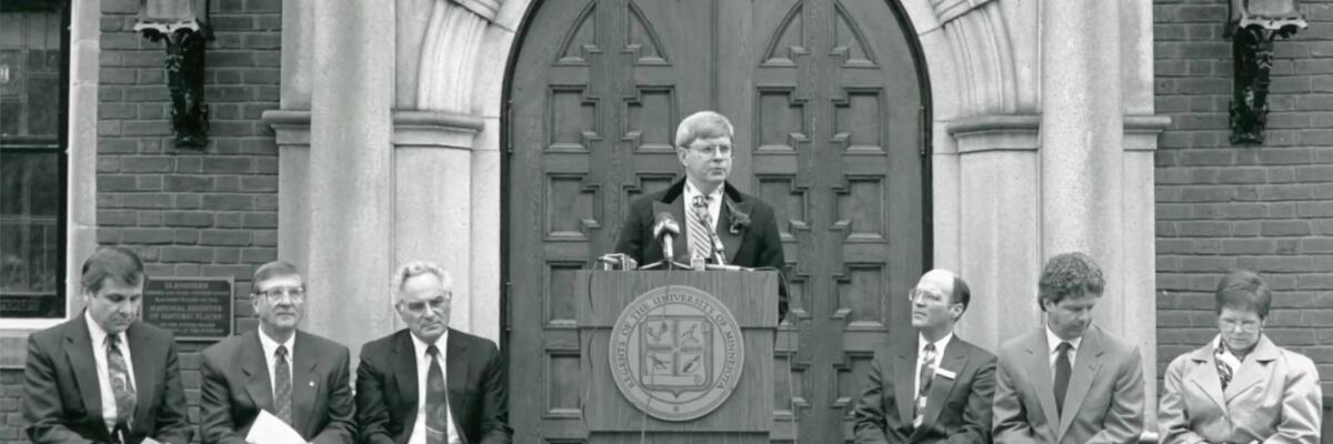 A man speaking in front of large mansion doors, six others seated on the sides.