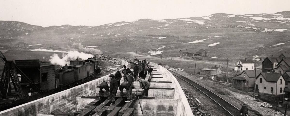 Men working on a railroad, hills in the background and a few small houses to the right.