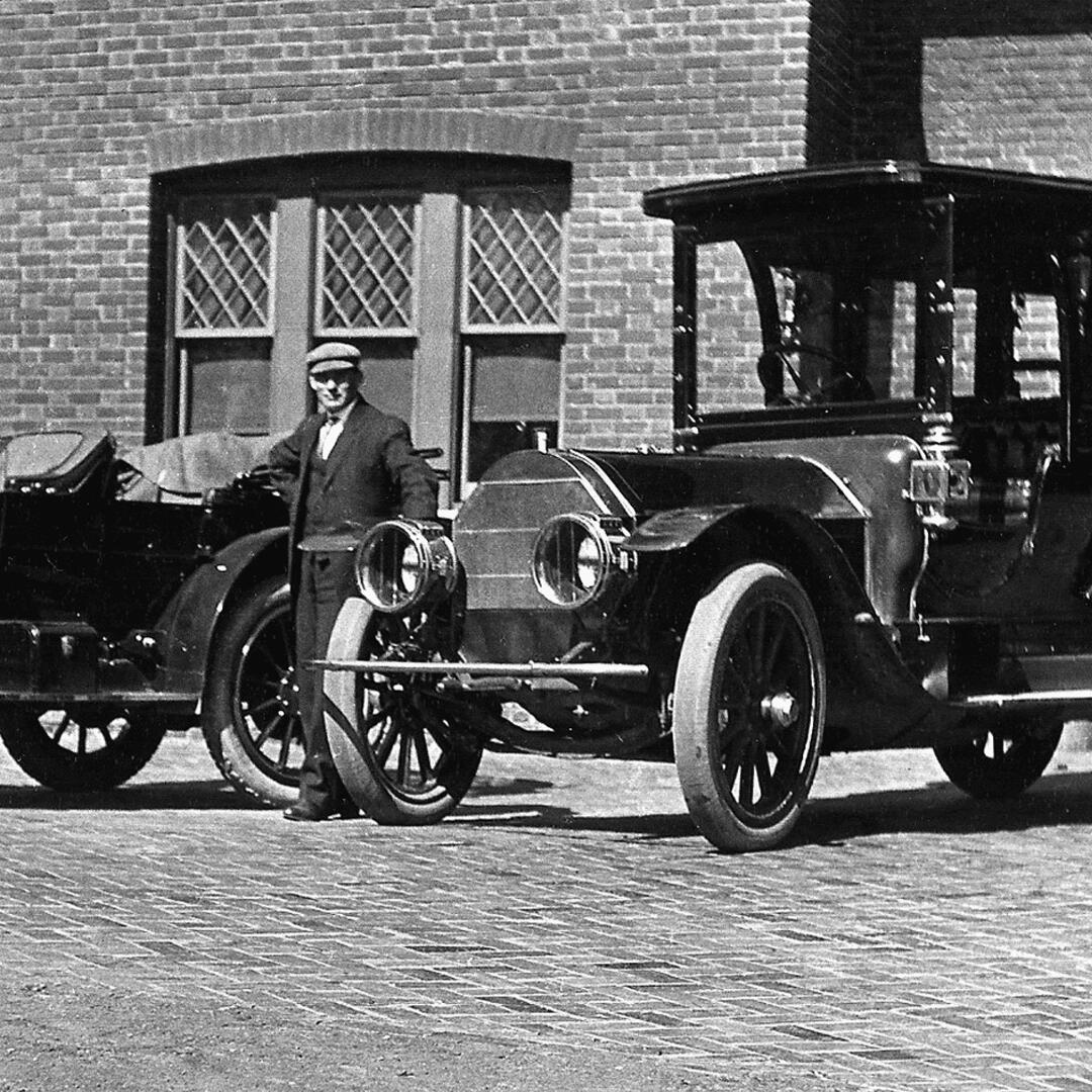 Chauffeur Rudolph Miller in front of the brick carriage house and standing by two vehicles.