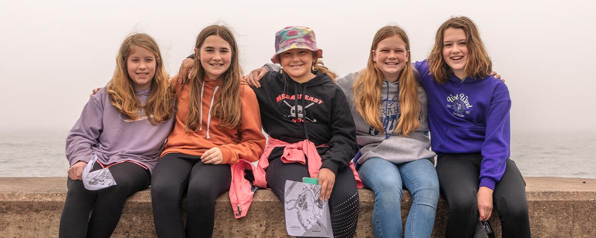 group of 5 girls smiling and sitting on the ledge of the concrete pier over Lake Superior.