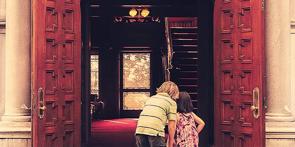 Two young kids looking through open front wood doors to the mansion and peering in.