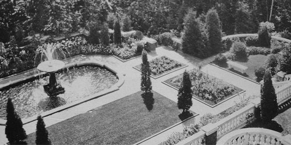 large marble fountain with a lawn of grass and pruned trees in front, brick paths in between.