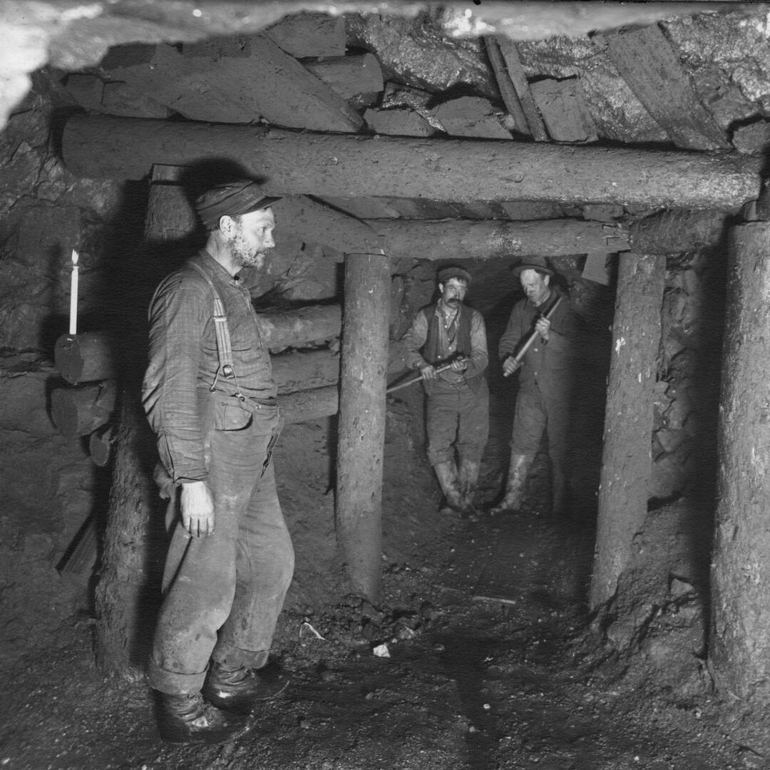 Three men underground with wood timbers and stone holding up the walls, two men holding pick axes for mining.