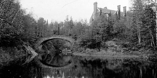 large opening of water (Tischer Creek) with a stone arch bridge above, surrounded by trees and a mansion in the background.