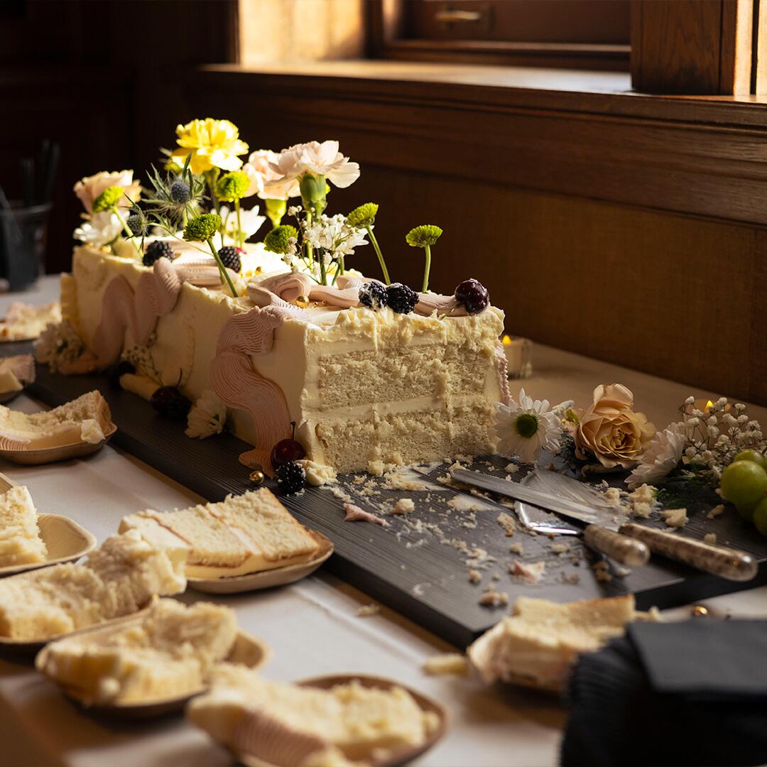long iced cake with swirls and flowers, cut on a table with pieces ready to take and eat.