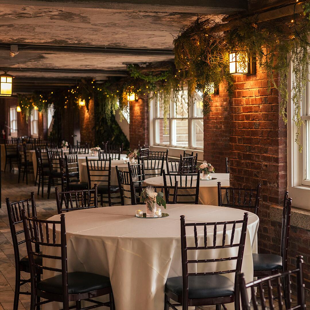 long brick room with greenery and lanterns near ceiling and round white tables with chairs and centerpieces.