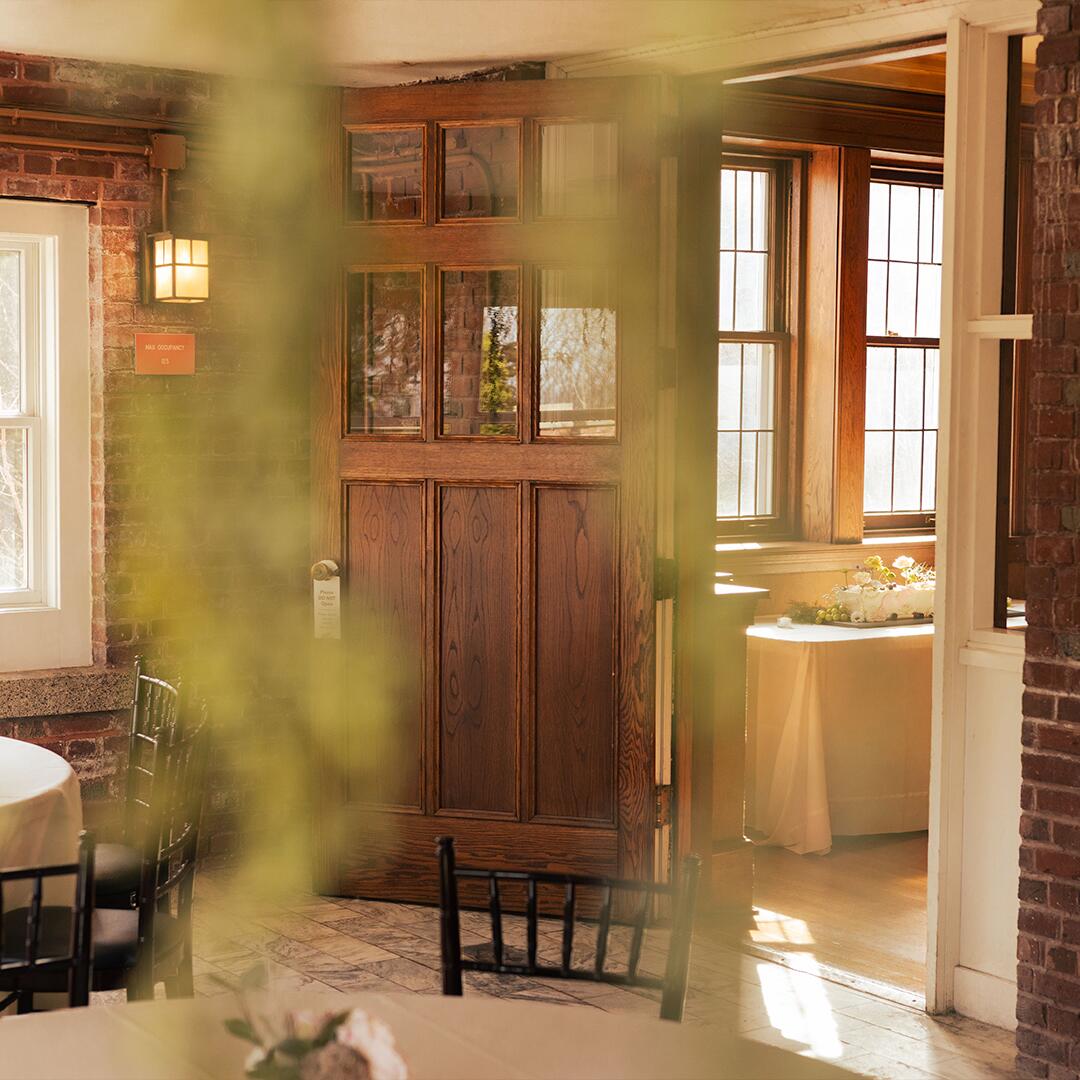 blurred hanging greenery in front of a room with round tables, an open door leading to room with a cake on the table.