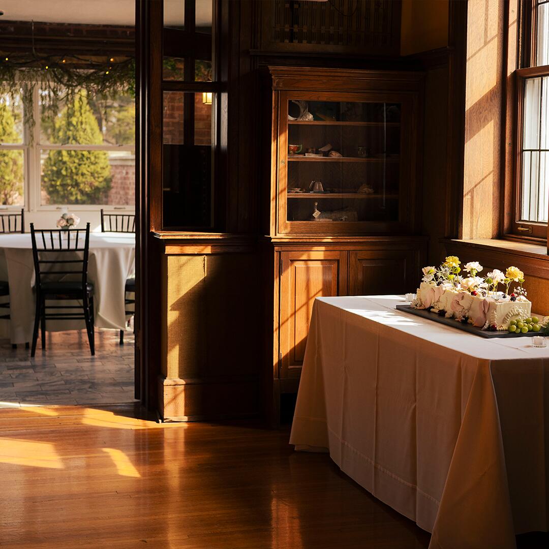 wood paneled room with a cake table and open door leading to a room with round tables and chairs.