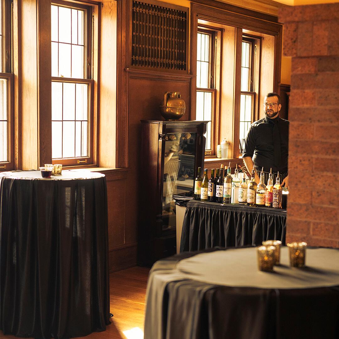 black cocktail tables and a bartender standing behind table with liquor, ready for guests.