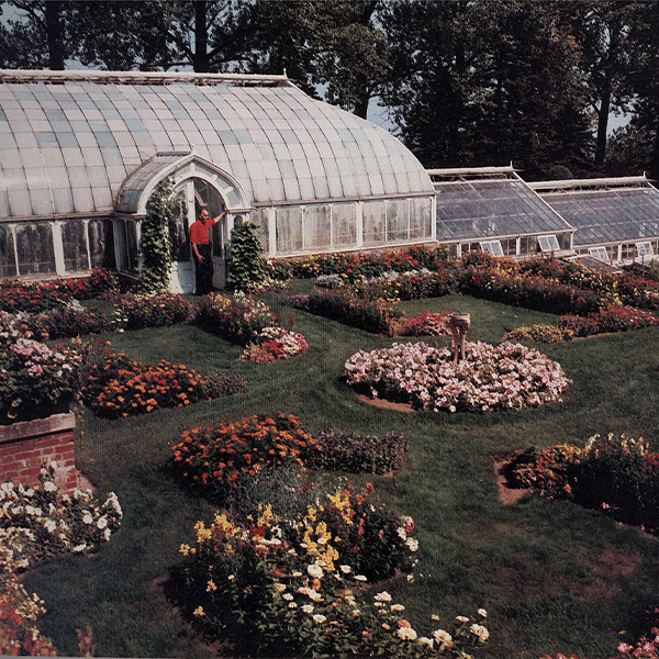 gardener standing in front of glass greenhouse and a yard of colorful flowers.