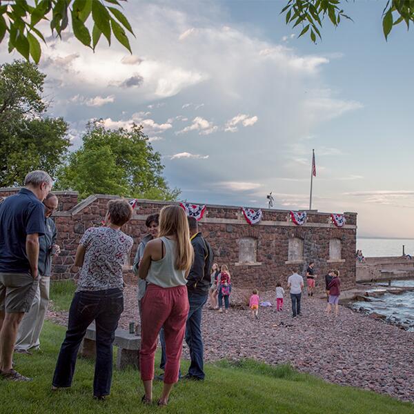 Adults talking and standing in a circle on the lawn, stone boathouse and pier in the background.