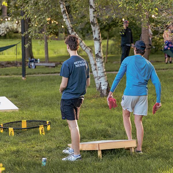 Two boys playing corn hole, throwing bean bags on the grass.