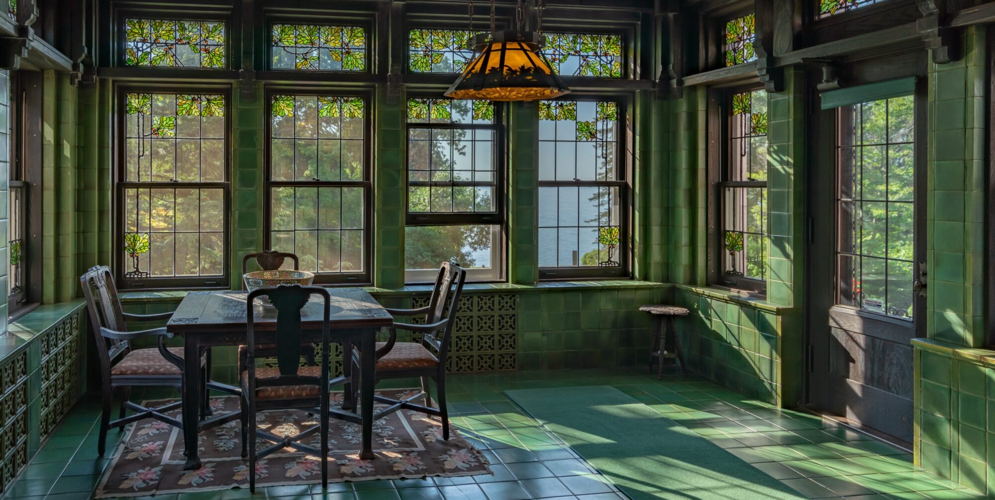 A green tiled room with a table in the corner and chairs around it. There is an orange light fixture hanging from the ceiling and one of the stained glass windows is ajar.