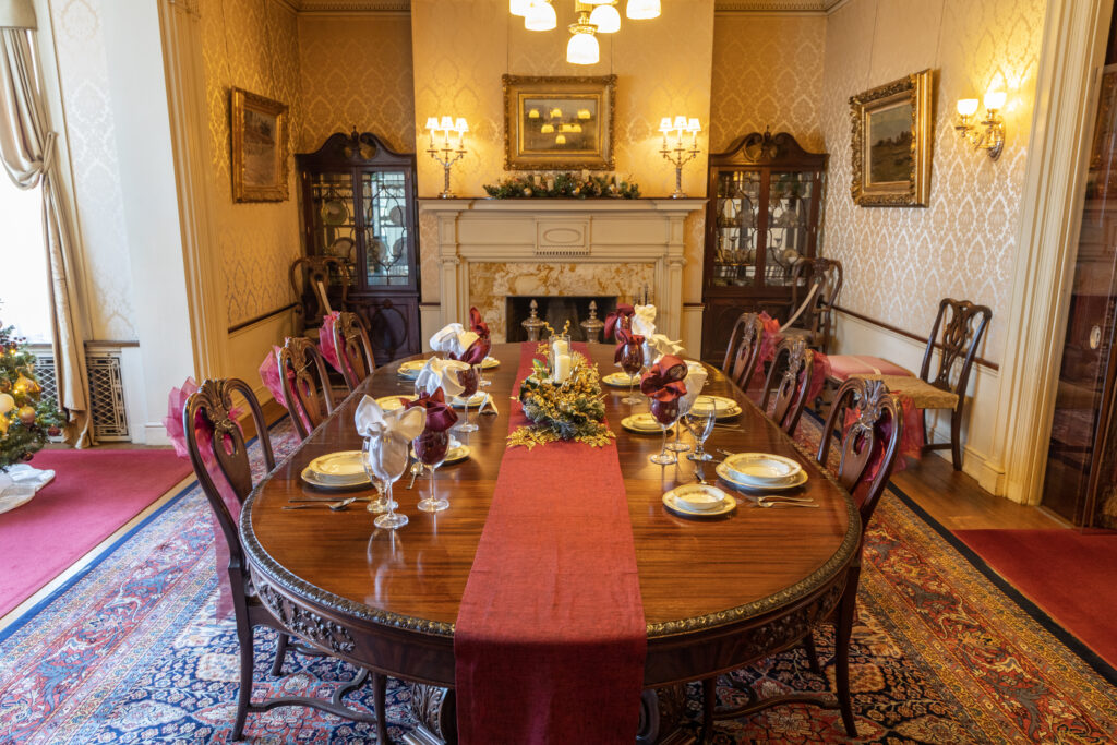A dining room table with a red runner and white china plates sits in a room with a chandelier over it surrounded by chairs and sitting on a red and white carpet.