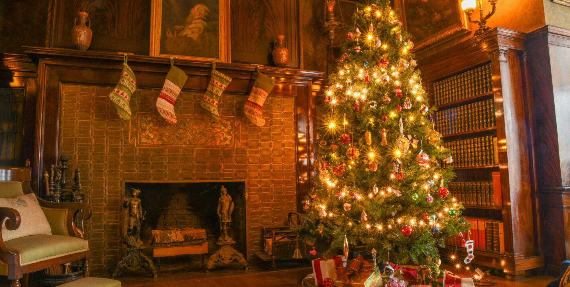 A Christmas tree stands next to a fireplace with stockings hung over it and a framed photo on top of it. There is a green sitting chair on the left of the photo.