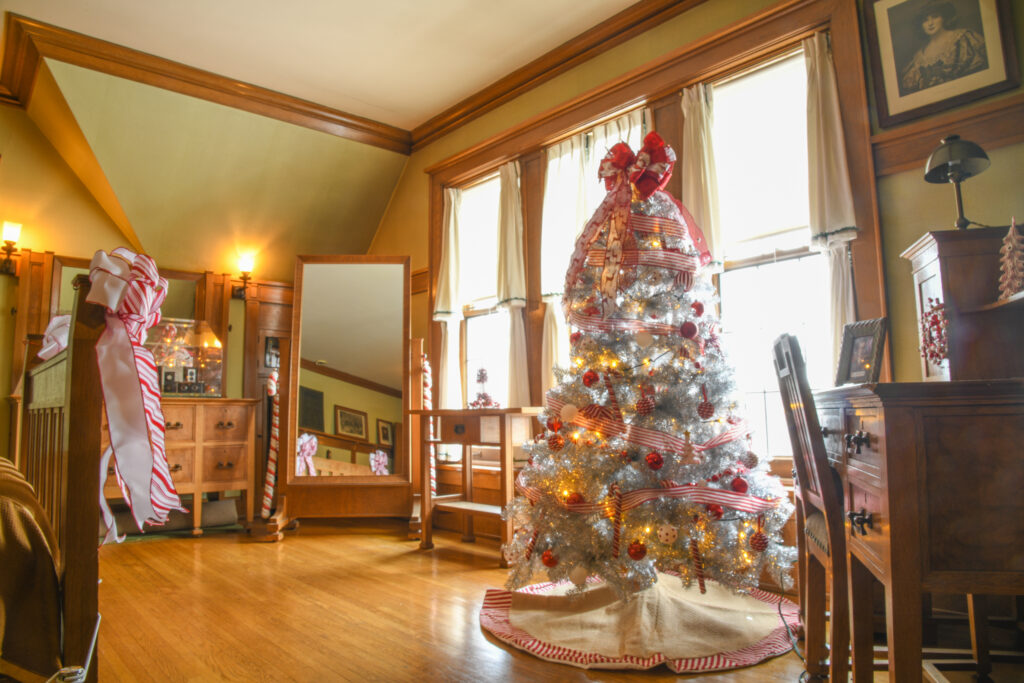 A candy cane colored tree stands lit and decorated on the right side of a room with a wooden desk on its right, bright windows behind it, wooden floors in front, and a mirror on the far side of the room.