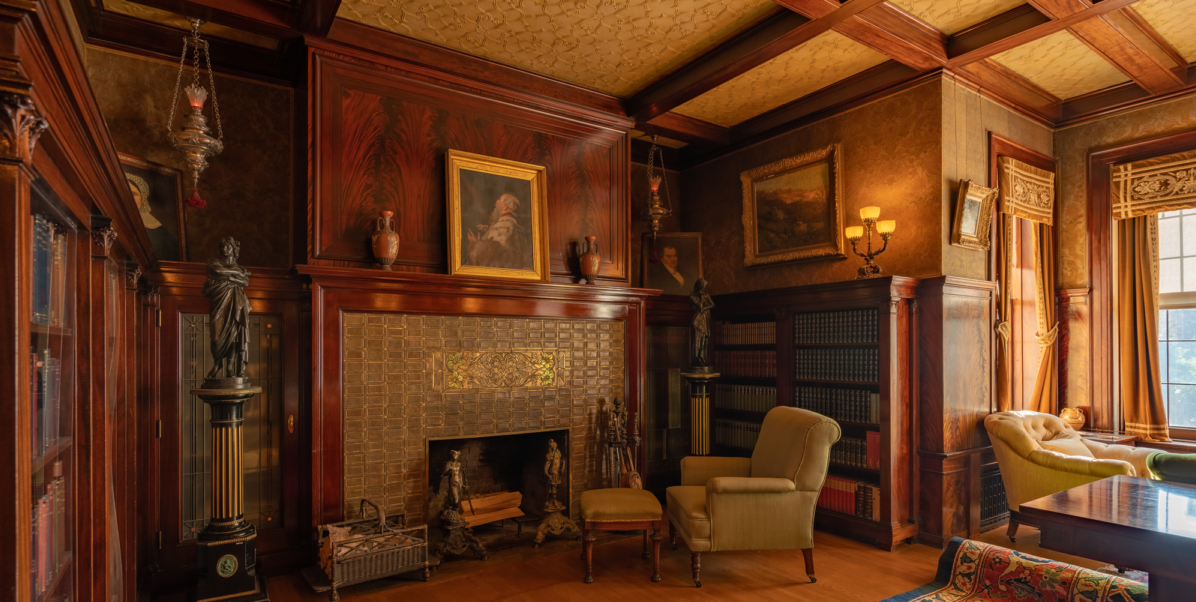 A picture of a library with a fireplace and a portrait on the wall with a chair facing the fireplace.
