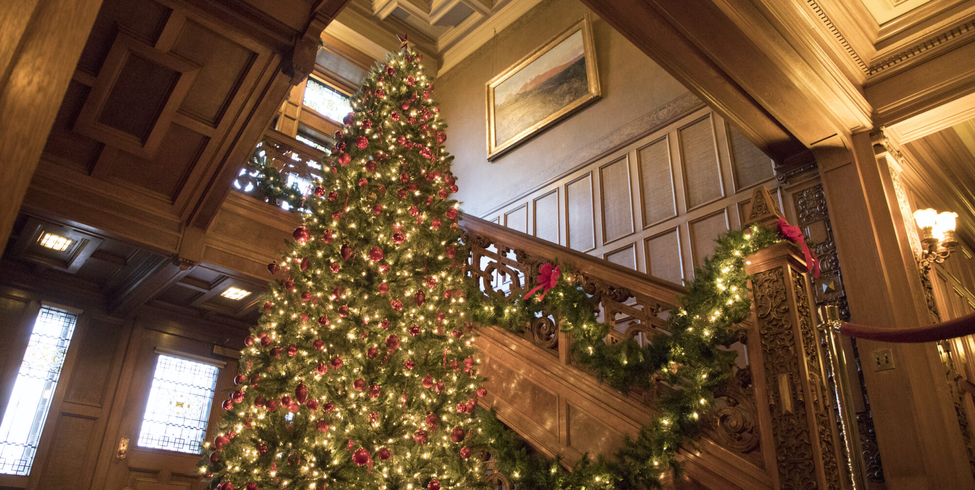 A tall Christmas tree lit with multi-color lights and ornaments stands in front of a wooden staircase and railing.